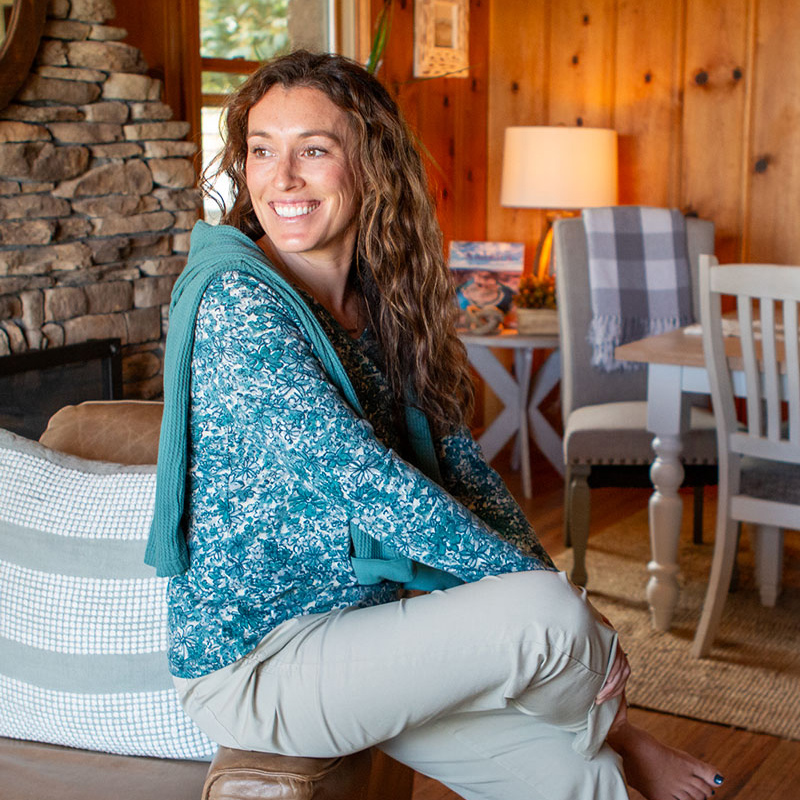 Woman sitting on arm of couch in wood cabin looking outdoors.